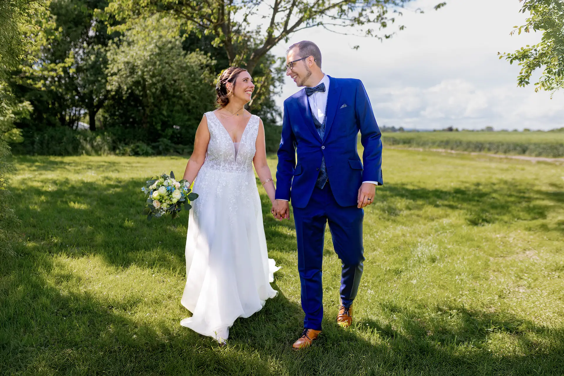 Braut und Bräutigam laufen Hand in Hand durch eine grüne Sommerwiese im goldenen Sonnenlicht