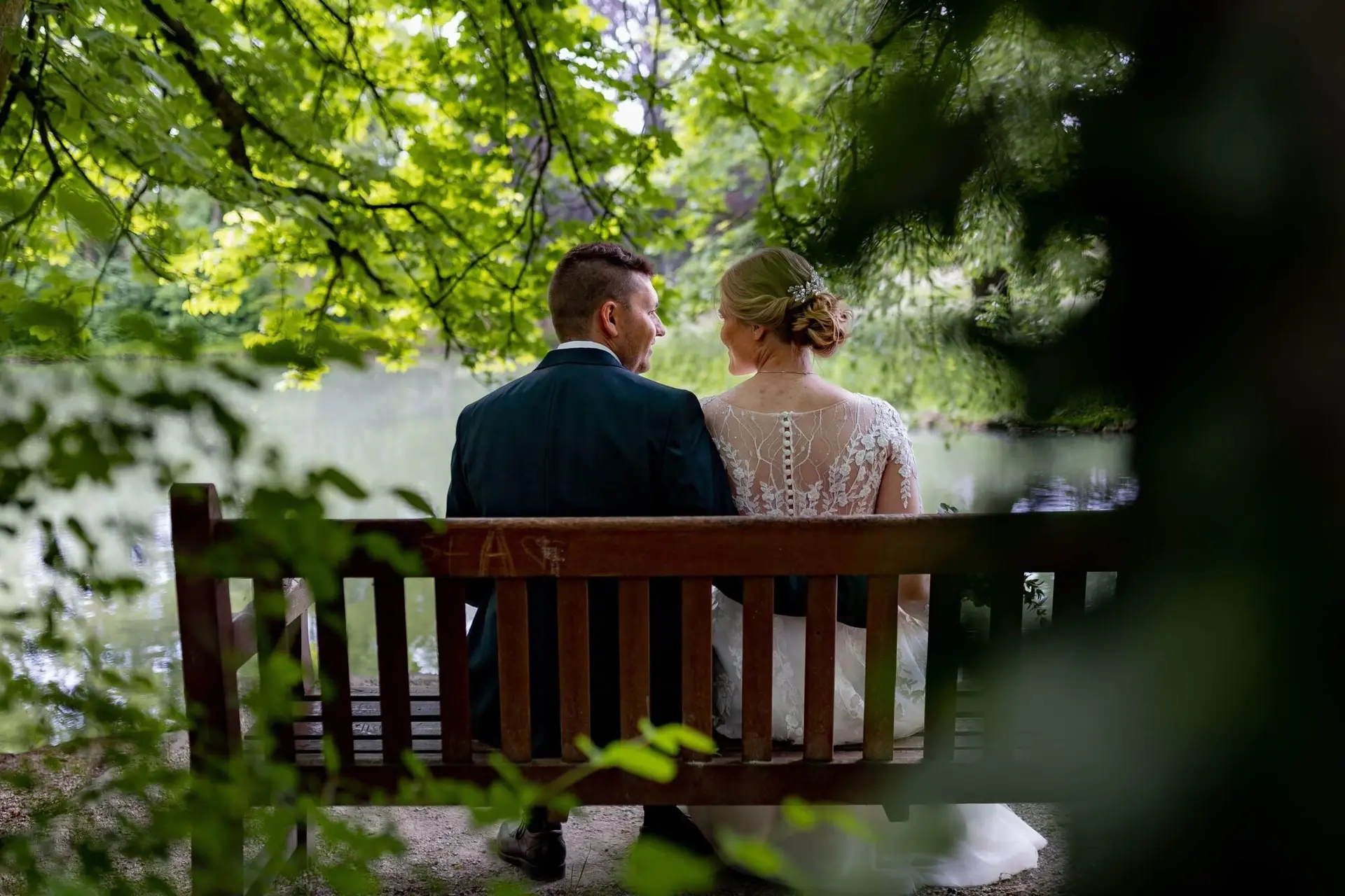 Brautpaar sitzt auf einer Bank am Teich im Schlossgarten Herrnsheim, Bräutigam im petrolfarbenen Anzug