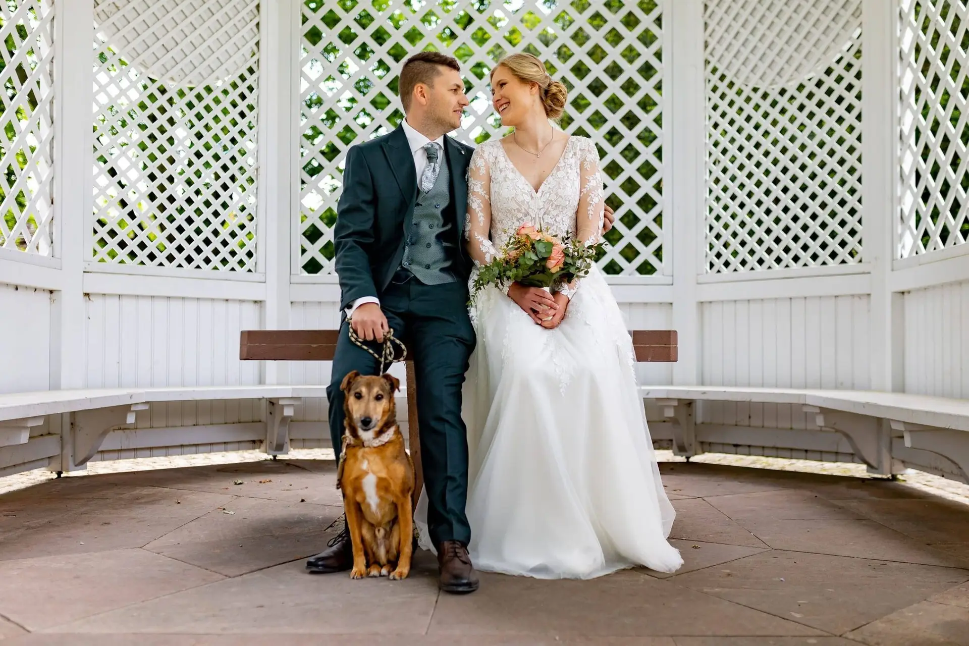 Brautpaar sitzt lächelnd mit ihrem braunen Hund in einer weißen Pergola im Schlossgarten Herrnsheim