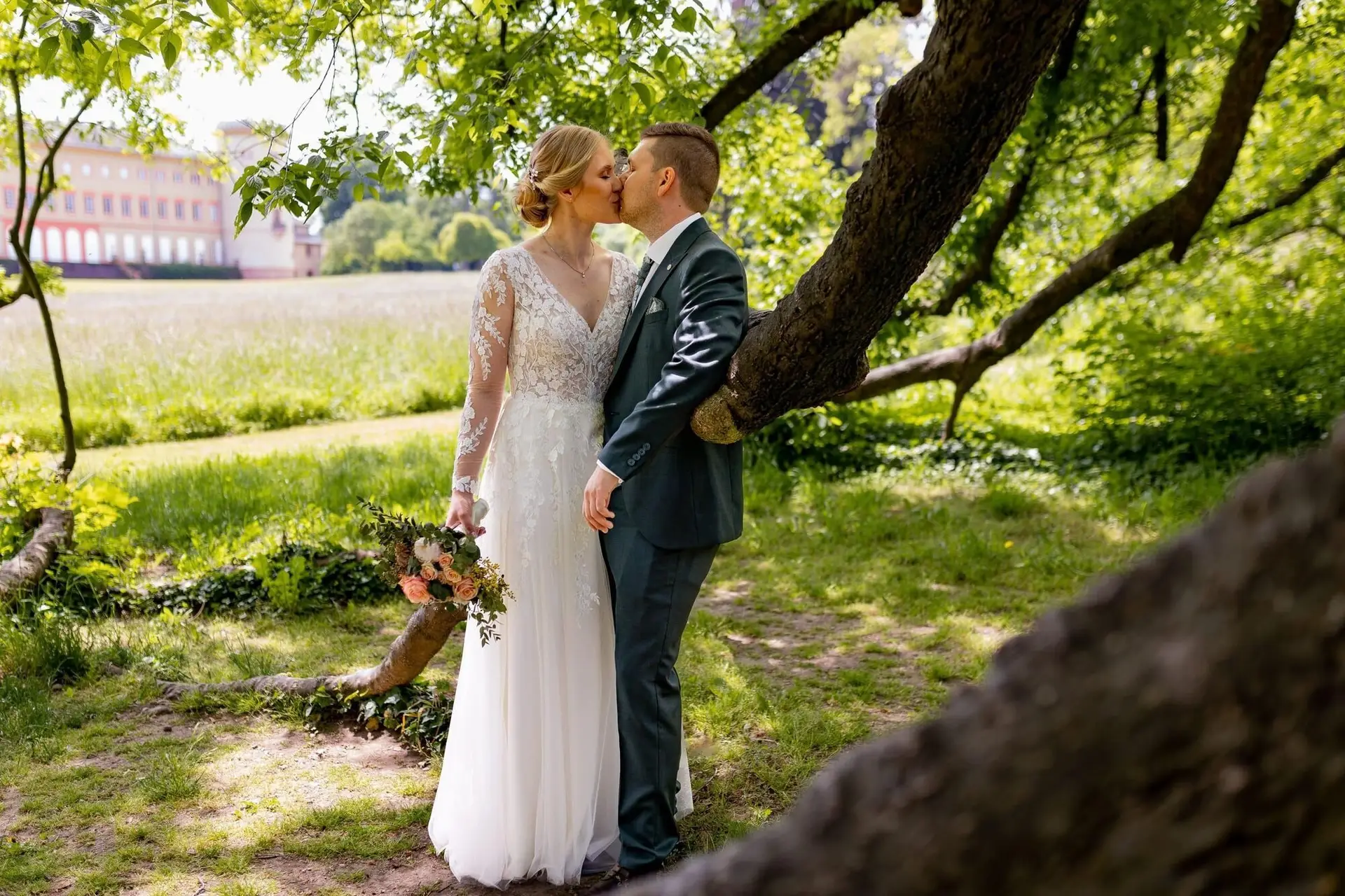 Brautpaar küsst sich leidenschaftlich unter einem alten Baum mit Schloss Herrnsheim im Hintergrund