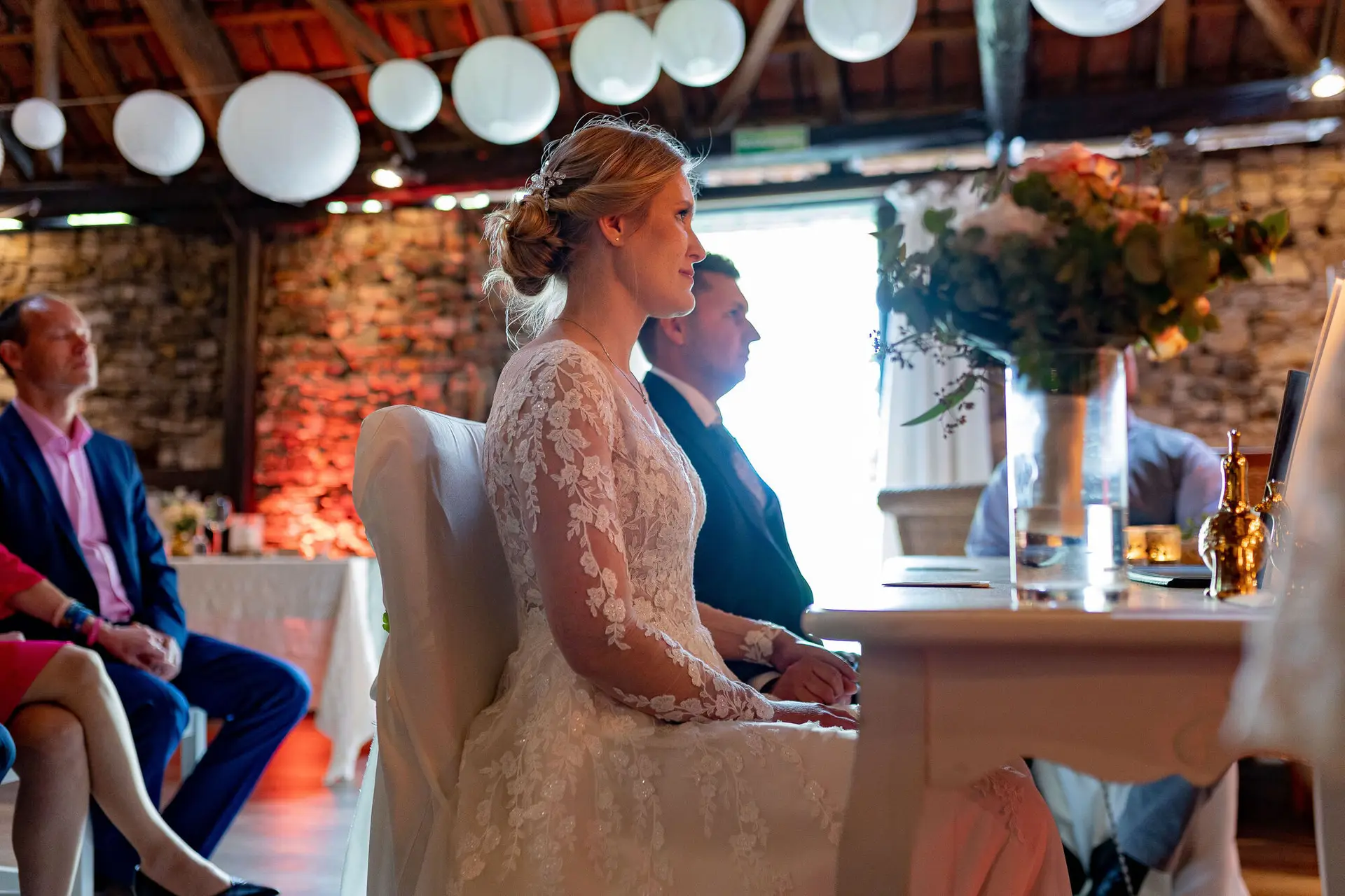 Blonde Braut sitzt an weißem Tisch unter hängenden weißen Laternen in historischem Steingemäuer