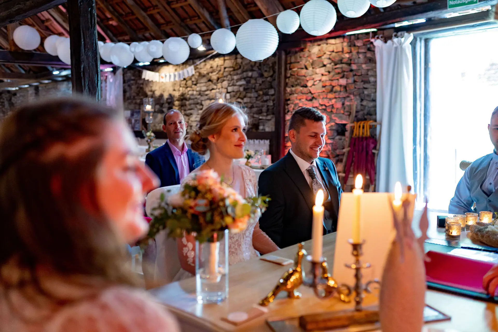 Brautpaar sitzt am langen Holztisch mit Gästen im rustikal dekorierten Festsaal mit weißen Laternen und warmem Kerzenlicht