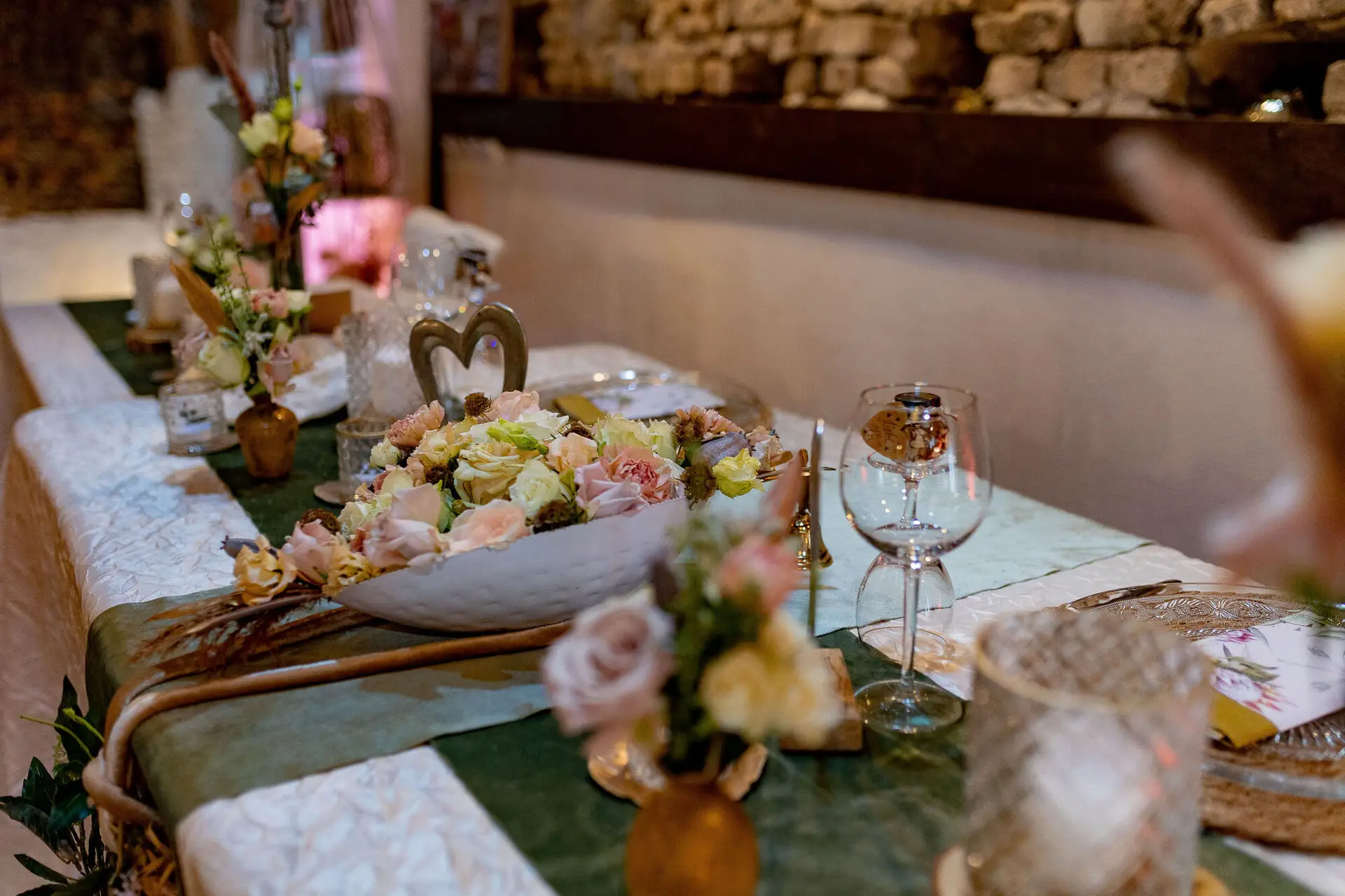 Langer Festtisch mit großer weißer Schale aus Pastellblüten und Kristallgläsern vor rustikaler Steinwand mit rosafarbener Beleuchtung